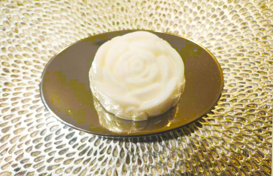 A white, rose-shaped mooncake sits gracefully on a reflective, oval-shaped dish with a textured background, evoking the stress-relief qualities of Dirty Soap Shoppe's White Rose Gardenia Flower Soap.