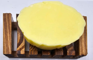 A round bar of Dirty Soap Shoppe's Yellow Rose Honeysuckle & Jasmine Flower Soap sits on a wooden soap dish with slats, offering stress relief and effective in reducing dark spots & acne.