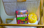 A box labeled "Dirty Soap Shoppe" sits beside a Yellow Rose Honeysuckle & Jasmine Flower Soap bar, both placed on a wicker basket, offering stress relief and reducing dark spots & acne.