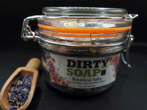 A glass jar with a label that reads "Dirty Soap Blue Cornflower and Calendula Flower" beside a wooden scoop with dried lavender.