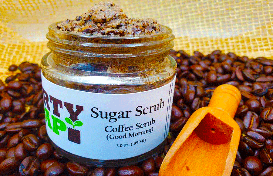A jar of Good Morning coffee sugar scrub on a bed of coffee beans with a wooden scoop by Dirty Soap Bubbles.