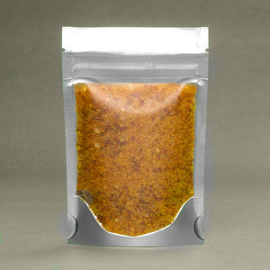 A sealed plastic pouch filled with brown granular substance and Burnt Orange & Turmeric Bath Salts from Dirty Soap on a neutral background.