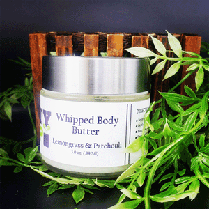 A jar of Lemongrass And Patchouli Whipped Body Butter from Dirty Soap Bubbles, surrounded by green leaves and a wooden holder, made with shea butter and cocoa butter.