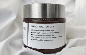 A jar of Dirty Soap Bubbles Mud Pie Sugar Scrub (Chocolate Lovers) with instructions on how to use printed on the label, resting on a draped white fabric.