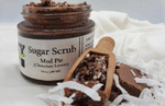 A jar of "Mud Pie Sugar Scrub (Chocolate Lovers)" organic sugar scrub with a wooden scoop and chocolate pieces from Dirty Soap Bubbles.