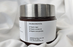 A jar of Mud Pie Sugar Scrub (Chocolate Lovers) by Dirty Soap Bubbles, listed among the ingredients, resting on a silky fabric.