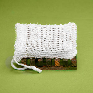 Crocheted white Dirty Soap soap saver bag resting on a small wooden pallet against a green background.